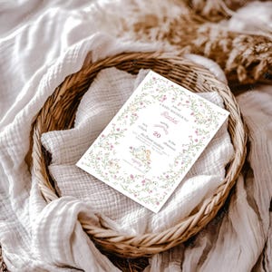 Puede incluir: Una invitaci&oacute;n para un baby shower con ilustraciones florales y de p&aacute;jaros, presentada en una cesta de mimbre. La invitaci&oacute;n presenta el texto "Baby Shower for Rachel" y los detalles del evento. El dise&ntilde;o incluye flores rosas y hojas verdes.
