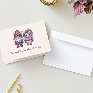 Könnte beinhalten: Eine Grußkarte mit rosa Hintergrund, auf der zwei Gnome in traditioneller schottischer Kleidung abgebildet sind. Der weibliche Gnome trägt ein rosa Kleid mit weißer Spitze und eine Blumenkrone. Der männliche Gnome trägt einen rot-grünen karierten Kilt. Der Text auf der Karte lautet "You and Me Are Meant To Be."