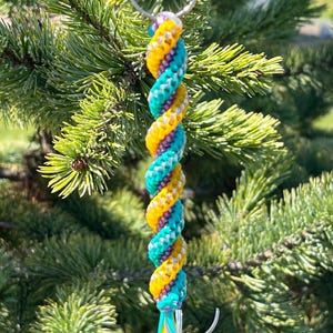 May include: A colorful keychain with a spiral design. The keychain features a twisted pattern of yellow, teal, and purple strands, attached to a silver metal ring. The keychain is hanging in front of a green, leafy background.