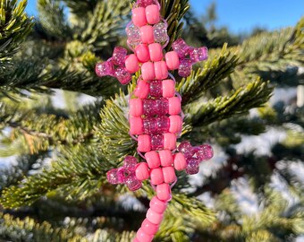 May include: A pink beaded lizard keychain hangs from a pine branch against a blue sky. The lizard is made of various shades of pink beads, with glittery accents on its legs and head. The keychain is a handmade accessory.