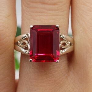 Può includere: Un anello d'argento con una grande pietra rossa di taglio smeraldo. L'anello ha un design delicato e ornato.