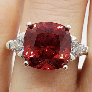 May include: A silver ring with a large, red, cushion-cut gemstone surrounded by three smaller, clear, round gemstones.