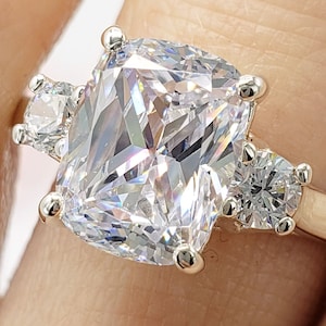 May include: A gold ring with a large, cushion-cut cubic zirconia center stone and two smaller round cubic zirconia accent stones.