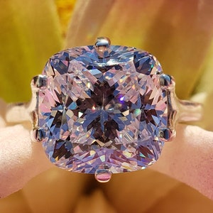 May include: A silver ring with a large, clear, cushion-cut gemstone. The ring is set against a pink and white flower.