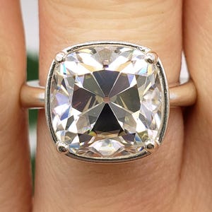 May include: A close-up of a silver ring featuring a large, square-cut, clear gemstone. The gemstone is faceted, reflecting light. The ring is set in a silver band. The initials "BB" are at the bottom of the image.