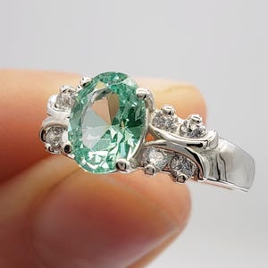 May include: A silver ring with a large oval green gemstone surrounded by smaller clear gemstones.