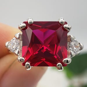 May include: A silver ring featuring a large, square-cut, deep red gemstone as the focal point. The ring is flanked by smaller, triangular, clear gemstones on each side. The initials "BB" are visible at the bottom.