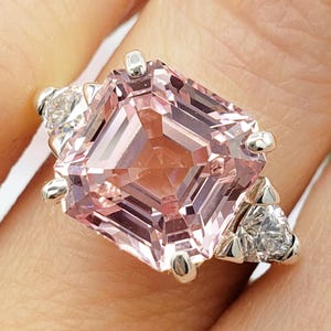 Puede incluir: Primer plano de un anillo con una gran gema rosa claro de talla cuadrada con gemas blancas triangulares m&aacute;s peque&ntilde;as a cada lado. El anillo est&aacute; engastado en una banda plateada. Las iniciales "BB" est&aacute;n en la parte inferior.