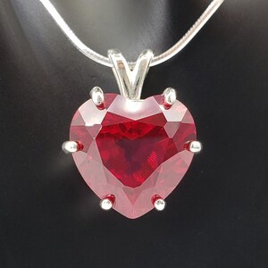 May include: A silver chain necklace with a heart-shaped pendant featuring a large, red gemstone. The pendant has a silver setting with small, decorative accents.