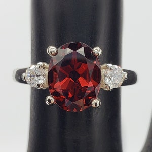 May include: A silver ring with a large oval-shaped red gemstone and two smaller round white gemstones on either side.