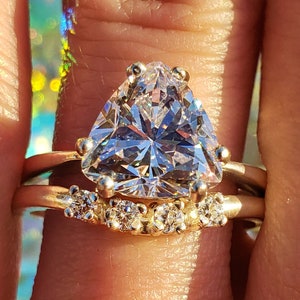 May include: A gold ring set with a large, clear, triangular-shaped gemstone. The ring is accented with smaller, clear gemstones on the band.
