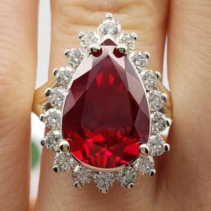 May include: A ring featuring a large, pear-shaped, deep red gemstone set in a metal band. The central stone is surrounded by a halo of numerous small, round, clear, sparkling stones. The letters BB are visible at the bottom of the image.