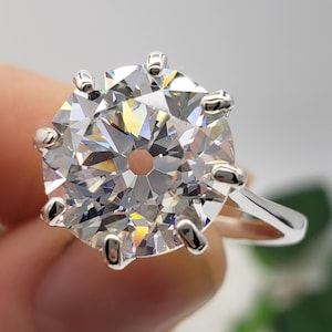 May include: A large, round-cut diamond ring with a silver band. The diamond is held in place by six prongs. The ring is held by a hand, with a blurred background.
