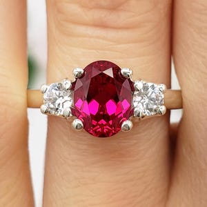 Könnte beinhalten: Ein goldener Ring mit einem großen, oval geschliffenen, tiefroten Edelstein, flankiert von zwei kleineren, runden, klaren Edelsteinen. Der Ring hat ein klassisches Drei-Stein-Design. Die Initialen "B.B" sind unten sichtbar.