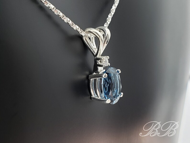 May include: A silver chain necklace with a pendant featuring a large oval-shaped blue topaz gemstone surrounded by smaller diamonds. The pendant has a delicate, intricate design.
