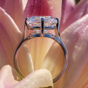 May include: A silver ring with a large, clear, round diamond set in a prong setting. The ring is resting on a pink flower petal.