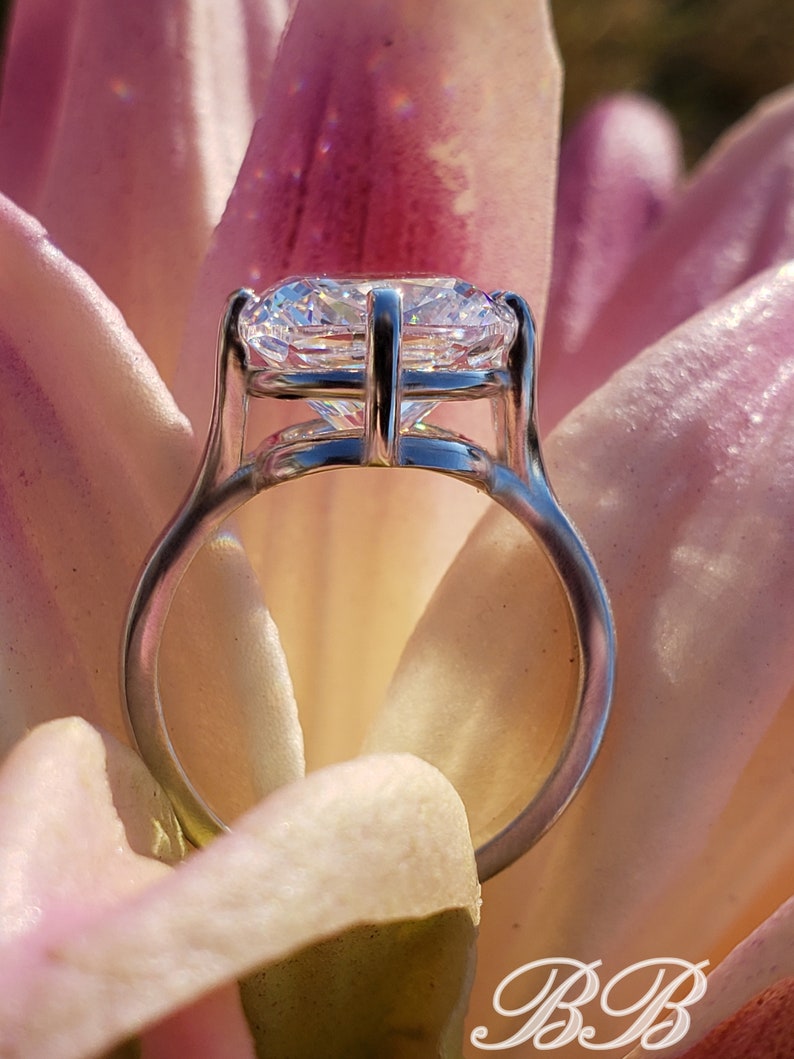 May include: A silver ring with a large, clear, round diamond set in a prong setting. The ring is resting on a pink flower petal.