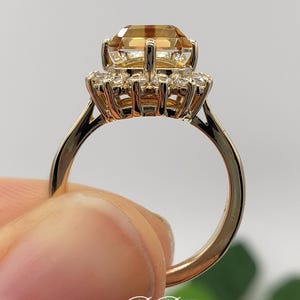 Pode incluir: Um anel de ouro com uma grande gema laranja de forma quadrada cercada por um halo de gemas brancas redondas menores.