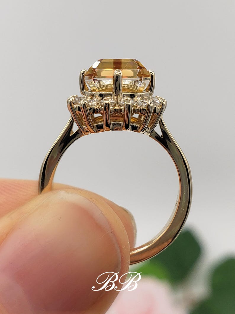 Pode incluir: Um anel de ouro com uma grande gema laranja de forma quadrada cercada por um halo de gemas brancas redondas menores.