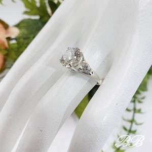 May include: A silver ring with a large, round, clear gemstone. The ring has intricate detailing on the band and around the central stone. The ring is displayed on a white surface with greenery in the background.