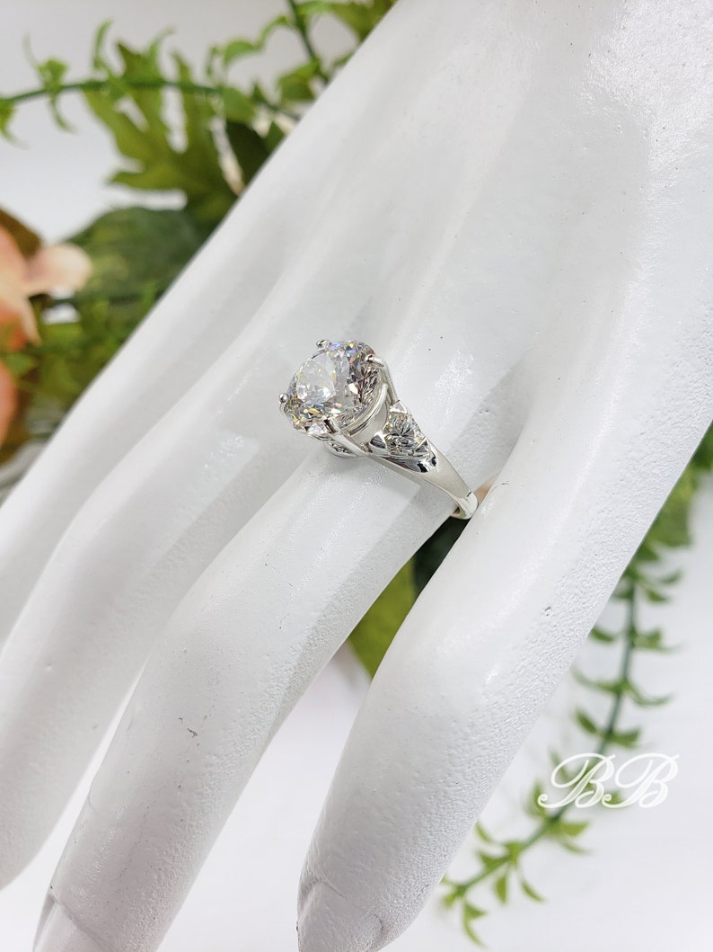May include: A silver ring with a large, round, clear gemstone. The ring has intricate detailing on the band and around the central stone. The ring is displayed on a white surface with greenery in the background.