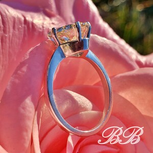 May include: A silver ring with a large, square-cut, light yellow gemstone. The ring is set against a soft pink rose, with the initials "BB" in a cursive script in the lower right corner.