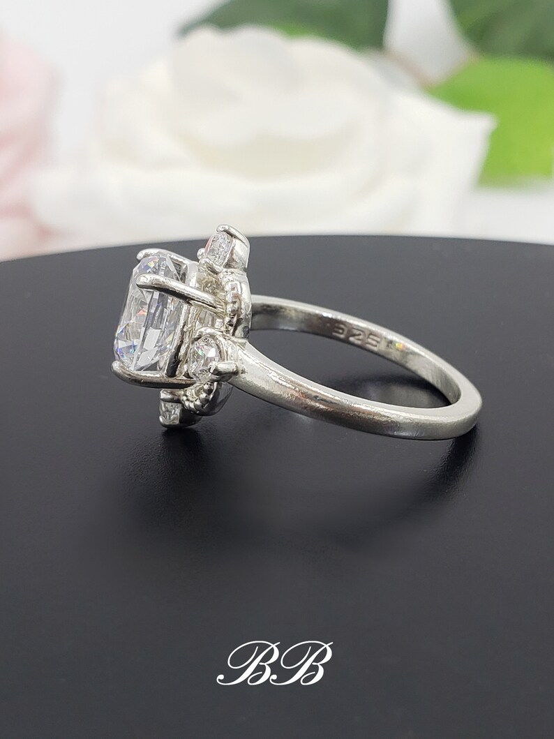 Puede incluir: Un anillo de plata con una piedra grande, transparente y ovalada rodeada de piedras m&aacute;s peque&ntilde;as y transparentes. El anillo est&aacute; sobre una superficie negra.