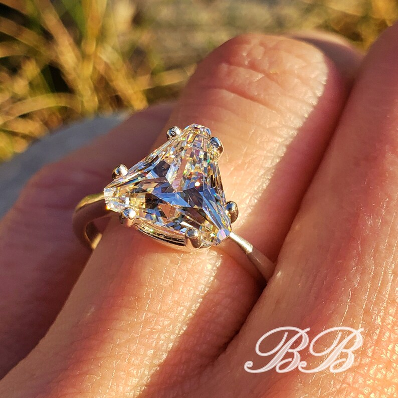 May include: A close-up of a silver ring featuring a large, triangular-cut, clear gemstone. The ring's band is thin and silver, with small prongs securing the gemstone. The ring is on a finger, with a blurred background.