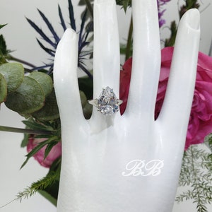 May include: A pear-shaped diamond engagement ring set in a silver band. The ring is on a white mannequin hand.