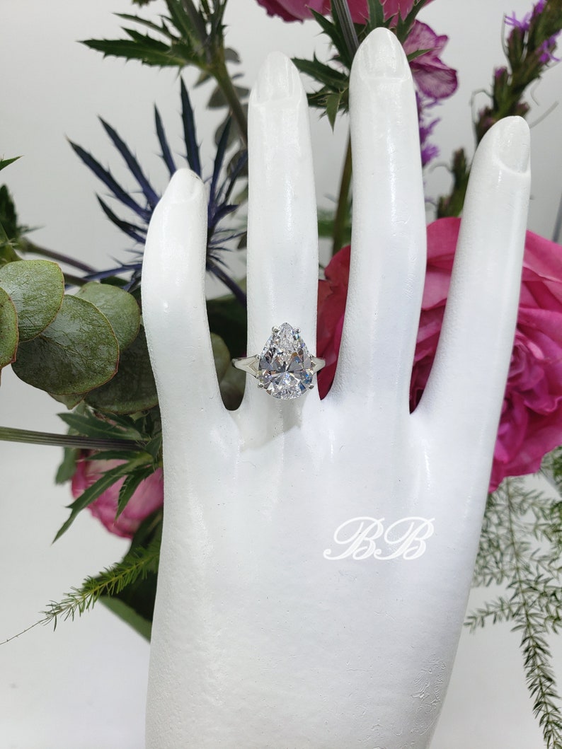 May include: A pear-shaped diamond engagement ring set in a silver band. The ring is on a white mannequin hand.