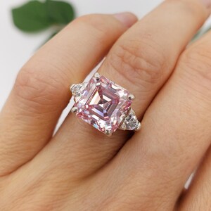 Puede incluir: Un anillo plateado con una gran gema rosa de corte cuadrado como pieza central, flanqueada por piedras claras triangulares m&aacute;s peque&ntilde;as. El anillo se muestra en una mano, mostrando el intrincado dise&ntilde;o y el brillo de la joya.