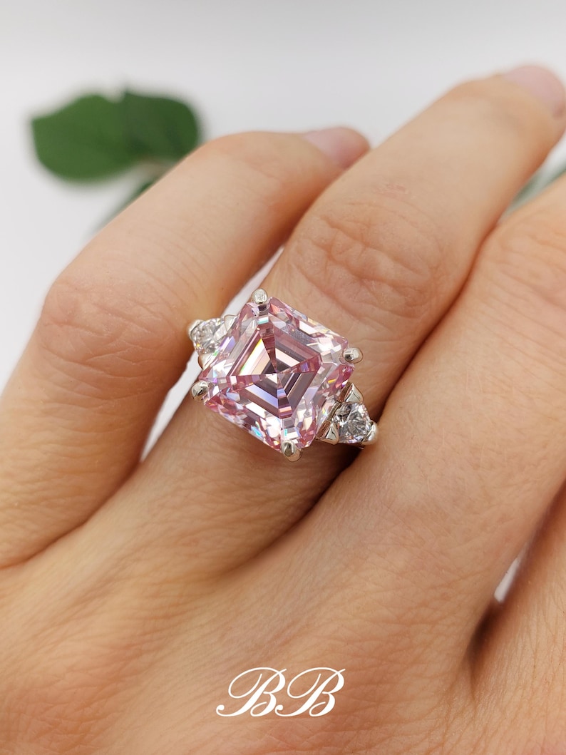 Puede incluir: Un anillo plateado con una gran gema rosa de corte cuadrado como pieza central, flanqueada por piedras claras triangulares m&aacute;s peque&ntilde;as. El anillo se muestra en una mano, mostrando el intrincado dise&ntilde;o y el brillo de la joya.