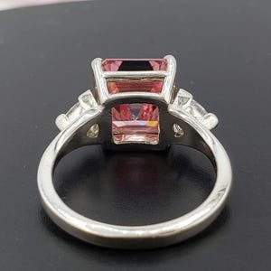 Puede incluir: Un anillo plateado con una gran gema rosa rectangular. Dos piedras triangulares m&aacute;s peque&ntilde;as flanquean la piedra principal. El anillo tiene una banda lisa y est&aacute; sobre un fondo oscuro. Las iniciales "BB" est&aacute;n en la parte inferior.
