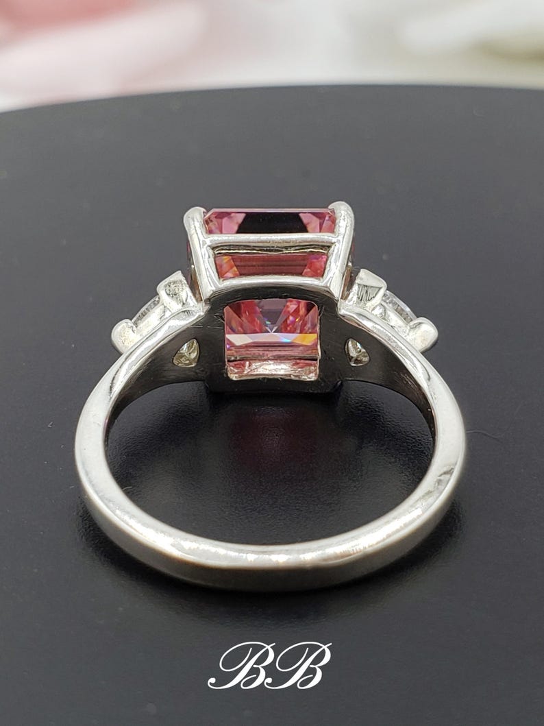 Puede incluir: Un anillo plateado con una gran gema rosa rectangular. Dos piedras triangulares m&aacute;s peque&ntilde;as flanquean la piedra principal. El anillo tiene una banda lisa y est&aacute; sobre un fondo oscuro. Las iniciales "BB" est&aacute;n en la parte inferior.