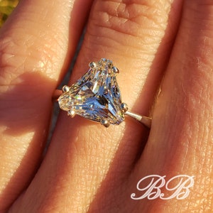 May include: A close-up of a silver ring featuring a large, clear, triangular-cut gemstone. The gemstone is faceted, reflecting light. The ring has small, gold-colored prongs holding the stone. The initials "BB" are in the lower right corner.