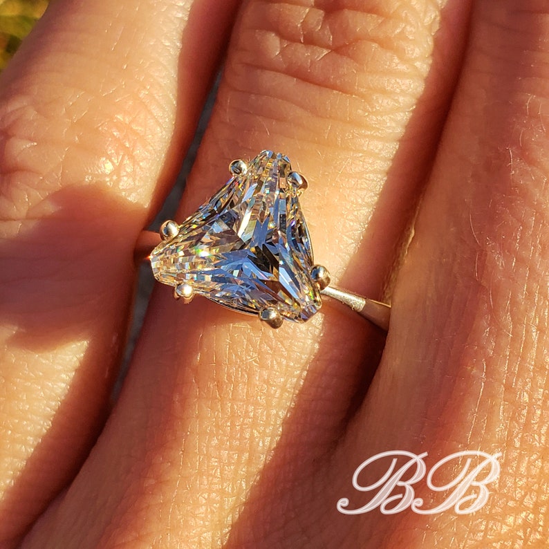 May include: A close-up of a silver ring featuring a large, clear, triangular-cut gemstone. The gemstone is faceted, reflecting light. The ring has small, gold-colored prongs holding the stone. The initials "BB" are in the lower right corner.