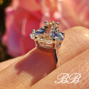 May include: Close-up of a silver ring with a large, triangular-cut gemstone. The gemstone is clear and faceted, catching the light. The ring is set against a soft pink background, with the initials "BB" in the lower right corner.