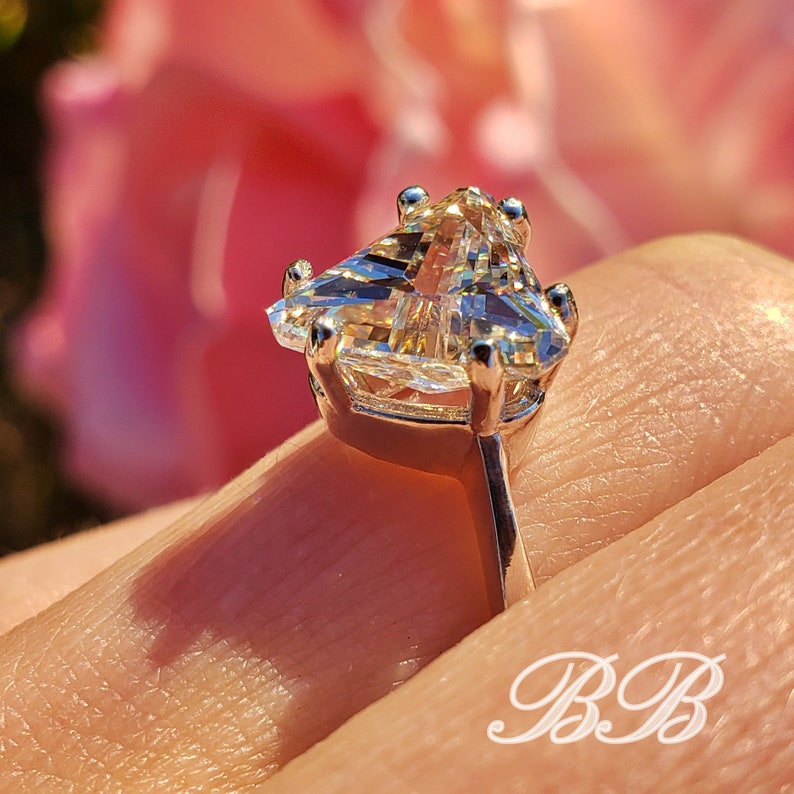 May include: Close-up of a silver ring with a large, triangular-cut gemstone. The gemstone is clear and faceted, catching the light. The ring is set against a soft pink background, with the initials "BB" in the lower right corner.