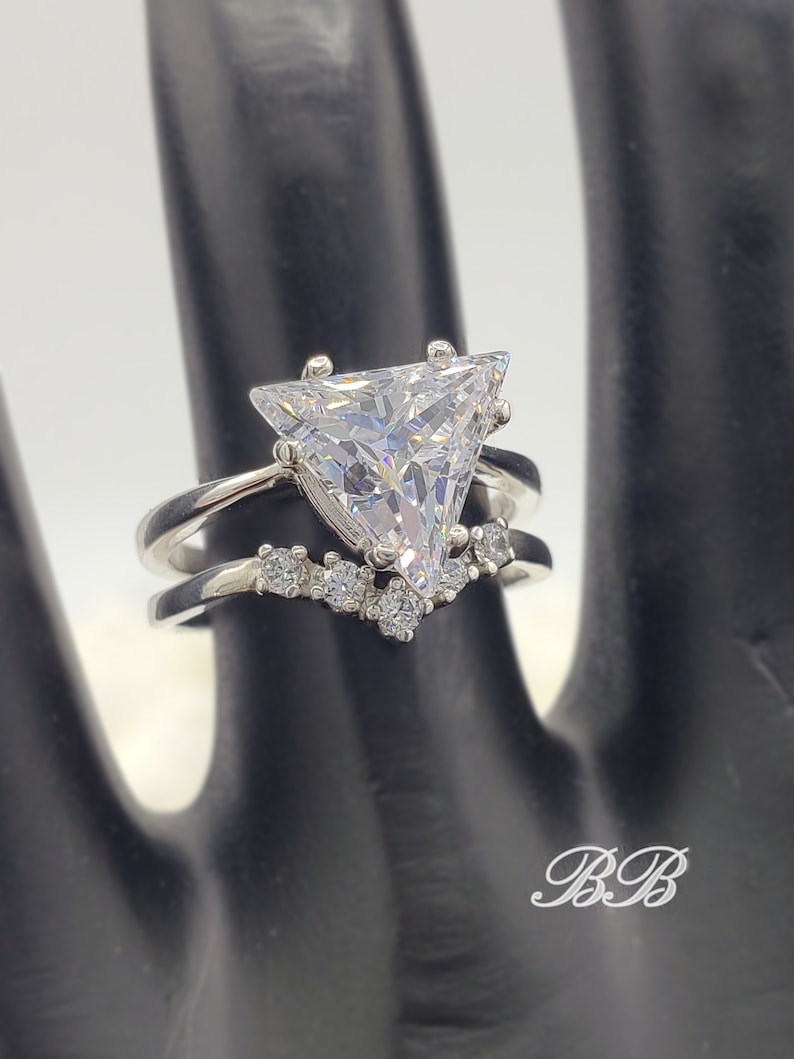 May include: A silver ring with a large triangular-shaped clear cubic zirconia stone. The ring has a smaller band with smaller clear cubic zirconia stones set in a v-shape.