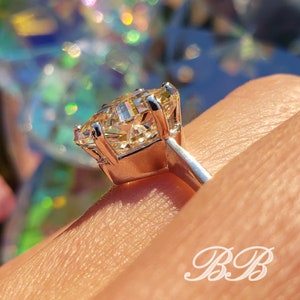 May include: A close-up of a silver ring with a large, radiant, yellow-toned gemstone. The gemstone is held in place by four prongs. The ring is on a finger, with the background out of focus.