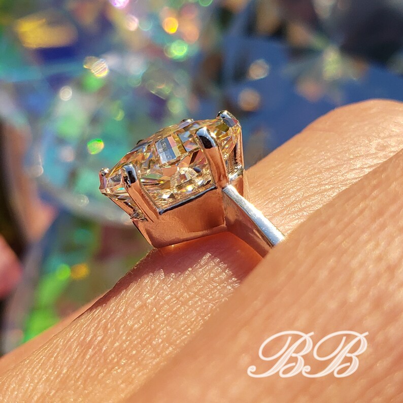 May include: A close-up of a silver ring with a large, radiant, yellow-toned gemstone. The gemstone is held in place by four prongs. The ring is on a finger, with the background out of focus.