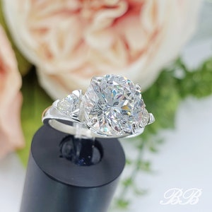 May include: A silver ring with a large, round, clear gemstone as the centerpiece, flanked by two smaller stones. The ring is set against a blurred floral background, with the initials "BB" in the lower right corner.