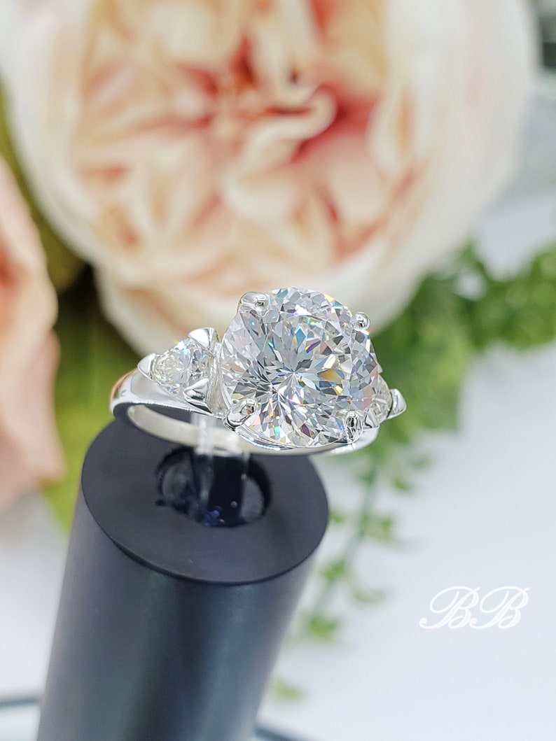 May include: A silver ring with a large, round, clear gemstone as the centerpiece, flanked by two smaller stones. The ring is set against a blurred floral background, with the initials "BB" in the lower right corner.