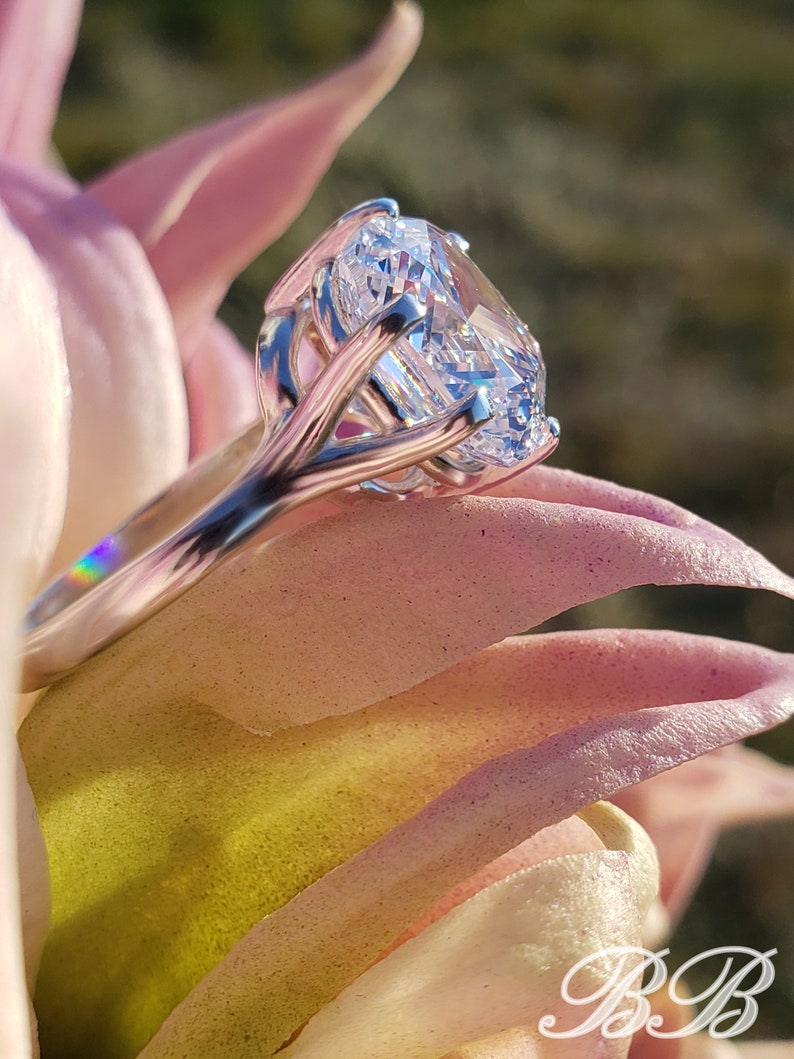 May include: A silver ring with a large, clear, round-cut diamond set in a prong setting. The ring is on a pink flower.