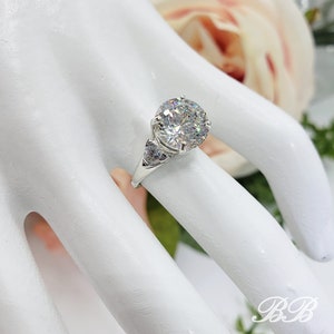 May include: A silver ring with a large, round, clear gemstone as the centerpiece. The ring has a classic design with smaller accent stones on the sides. The ring is displayed on a white surface with a soft, blurred floral background.