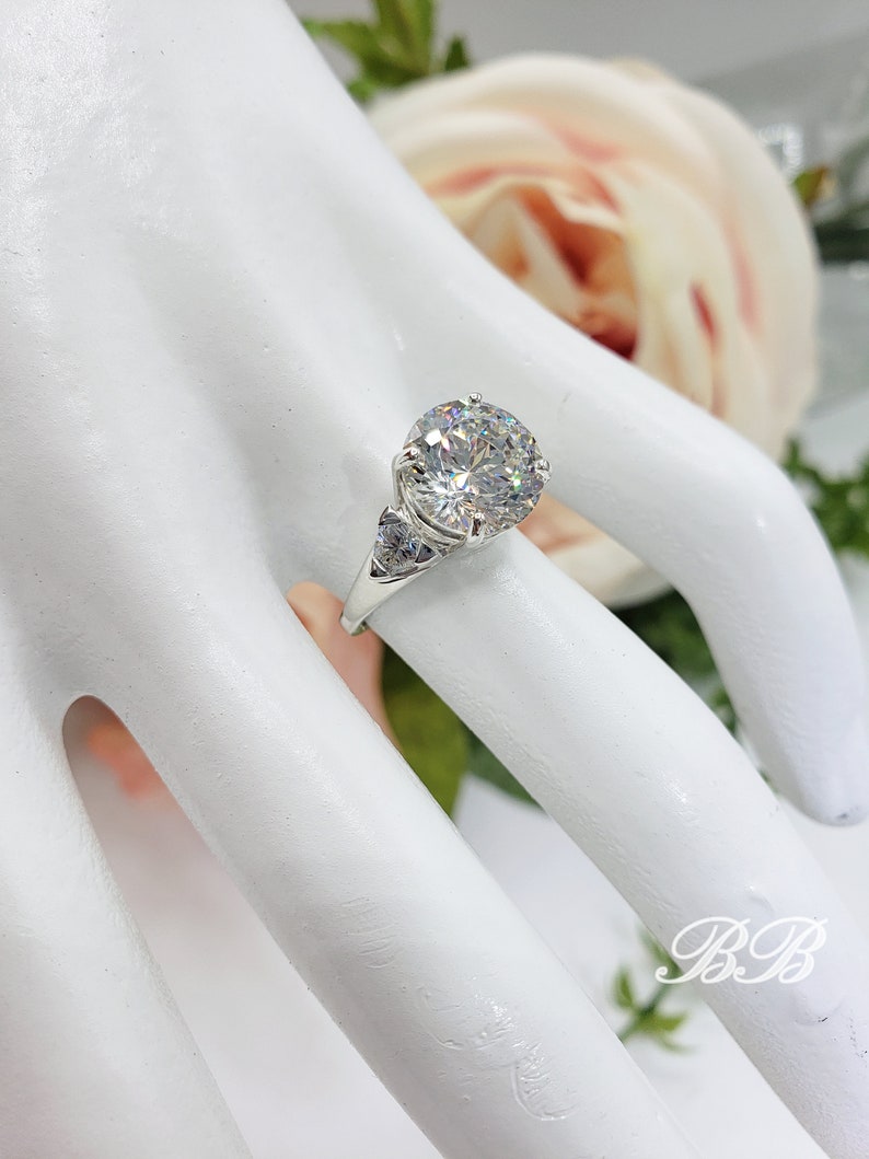 May include: A silver ring with a large, round, clear gemstone as the centerpiece. The ring has a classic design with smaller accent stones on the sides. The ring is displayed on a white surface with a soft, blurred floral background.