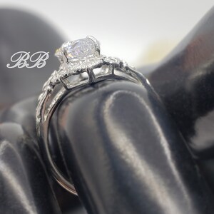 Puede incluir: Un anillo de plata con una gran piedra redonda transparente rodeada de piedras m&aacute;s peque&ntilde;as. El anillo est&aacute; en un expositor de anillos negro.