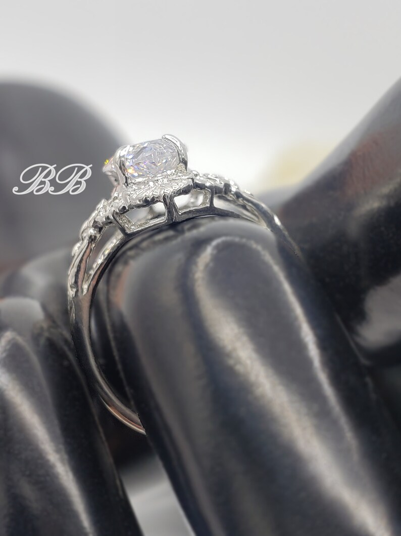 Puede incluir: Un anillo de plata con una gran piedra redonda transparente rodeada de piedras m&aacute;s peque&ntilde;as. El anillo est&aacute; en un expositor de anillos negro.