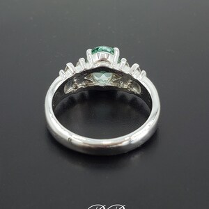 Puede incluir: Un anillo de plata con una piedra preciosa verde rodeada de piedras preciosas m&aacute;s peque&ntilde;as y transparentes. El anillo est&aacute; sobre una superficie negra.
