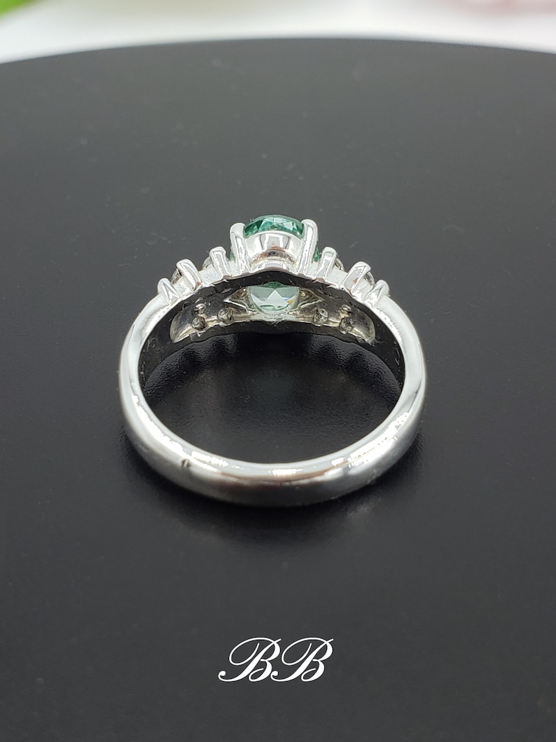 Puede incluir: Un anillo de plata con una piedra preciosa verde rodeada de piedras preciosas m&aacute;s peque&ntilde;as y transparentes. El anillo est&aacute; sobre una superficie negra.